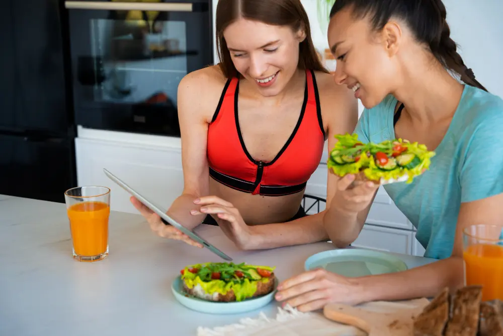 Duas mulheres jovens com roupas esportivas olhando tablet enquanto comem refeição low carb