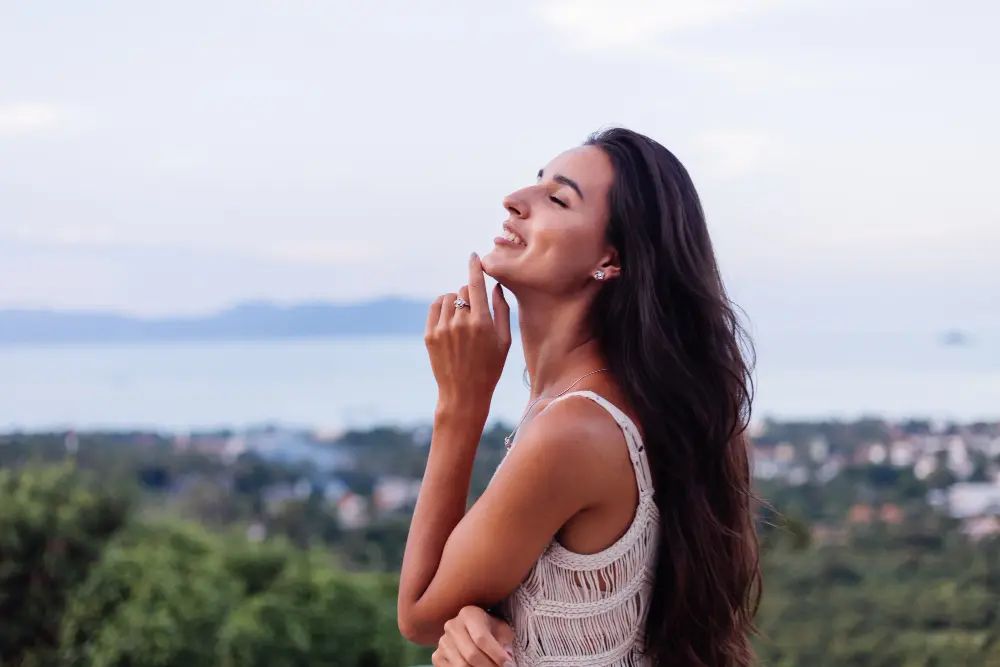 Mulher de lado com o dedo no queixo enquanto sorri de olhos fechados em fundo desfocado de natureza e cidade distante