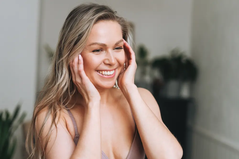 Mulher loira sorrindo com mãos nas bochechas em fundo doméstico com plantas desfocado