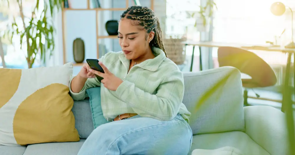 Mulher sentada em sofá segurando celular com sorriso enquanto faz compras online 