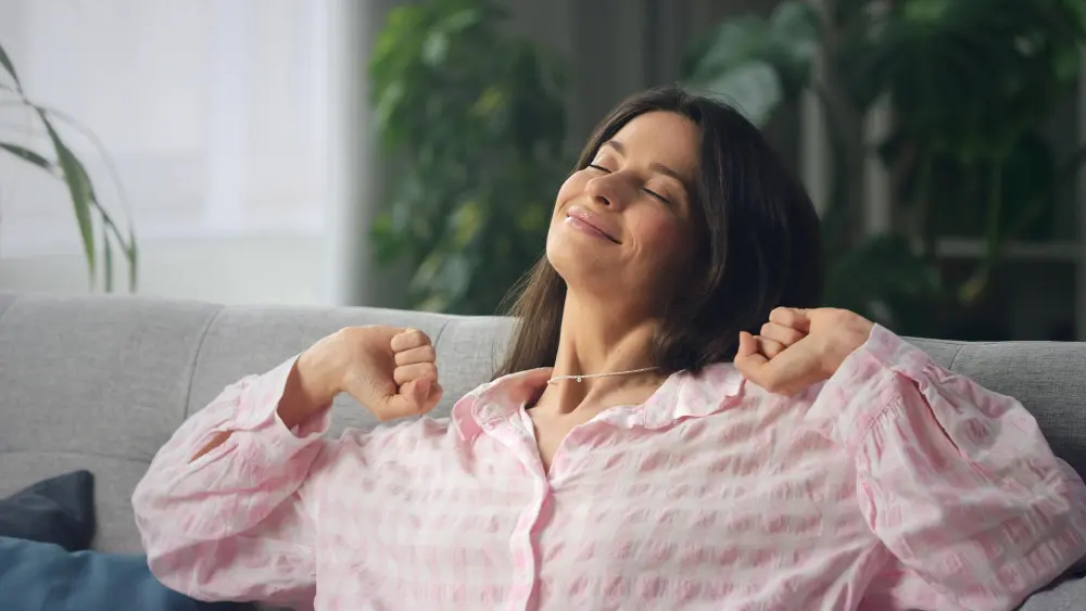 Mulher praticando mindfulness sentada em sofá sorrindo de olhos fechados com mãos para cima se espreguiçando.