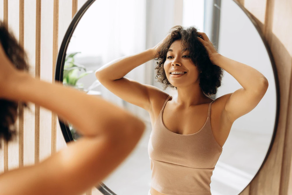 Mulher com pele saudável sorrindo no espelho com as mãos na cabeça