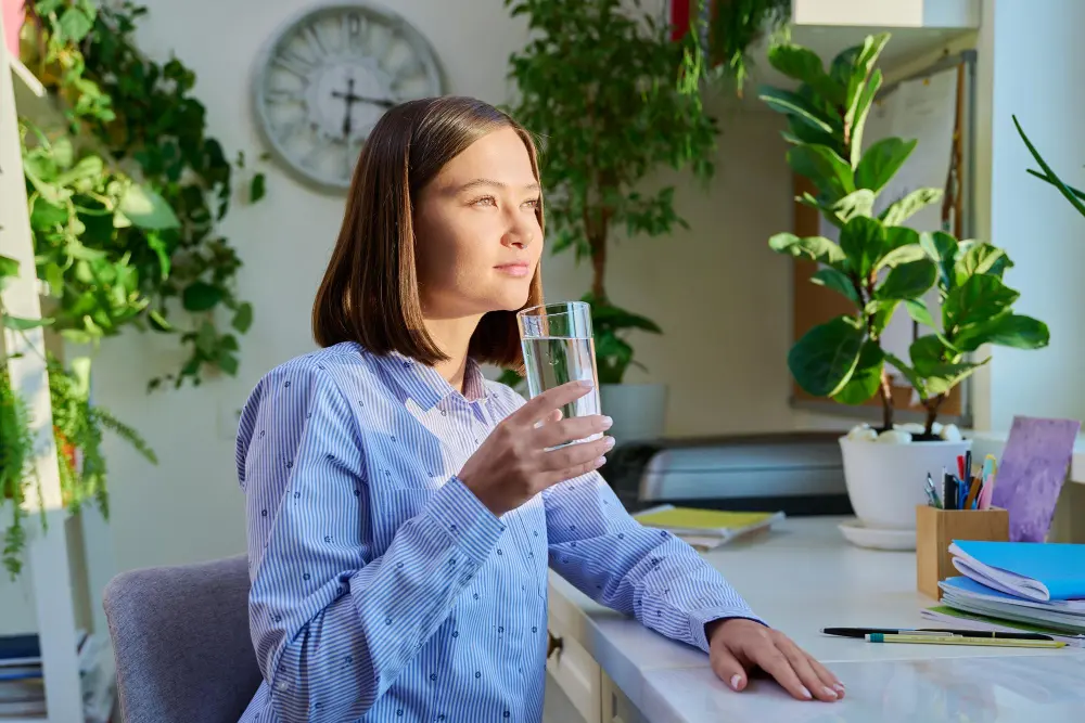 Mulher cuidando da Saúde e clareza mental com copo de água na mão sentada ao lado de plantas e iluminada pela luz natural solar
